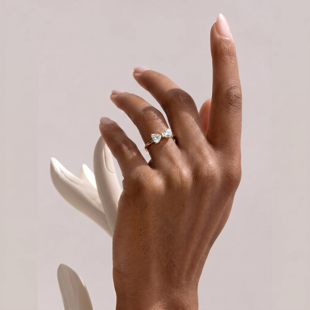 An elegant, dark-skinned hand gracefully displays the golden bow ring with heart-shaped clear stones. The hand is posed alongside a modern white sculptural element, emphasizing the jewelry's sophisticated appeal.