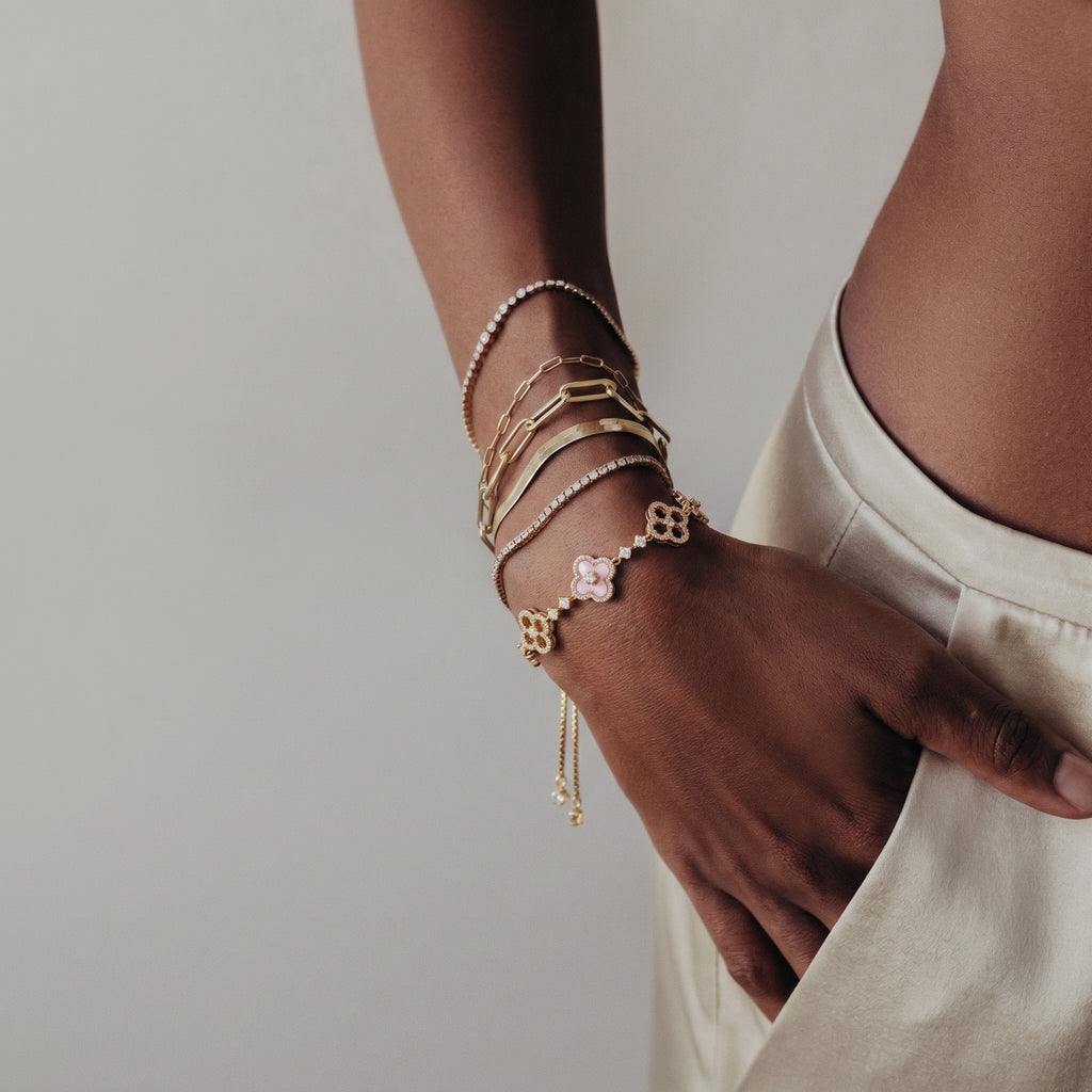 Woman's hand elegantly adorned with a stack of gold-toned bracelets, prominently featuring the Clover Bloom bolo bracelet with pink enamel-effect petals and simulated diamonds, against a minimalist backdrop.