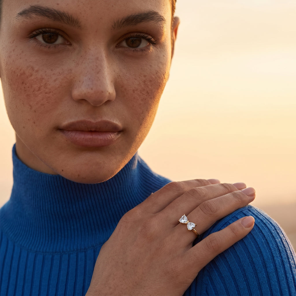 A woman with freckles models the golden bow ring featuring heart-shaped stones, elegantly resting her hand on her shoulder. The scene is bathed in warm, soft sunset light, highlighting the ring's refined beauty.