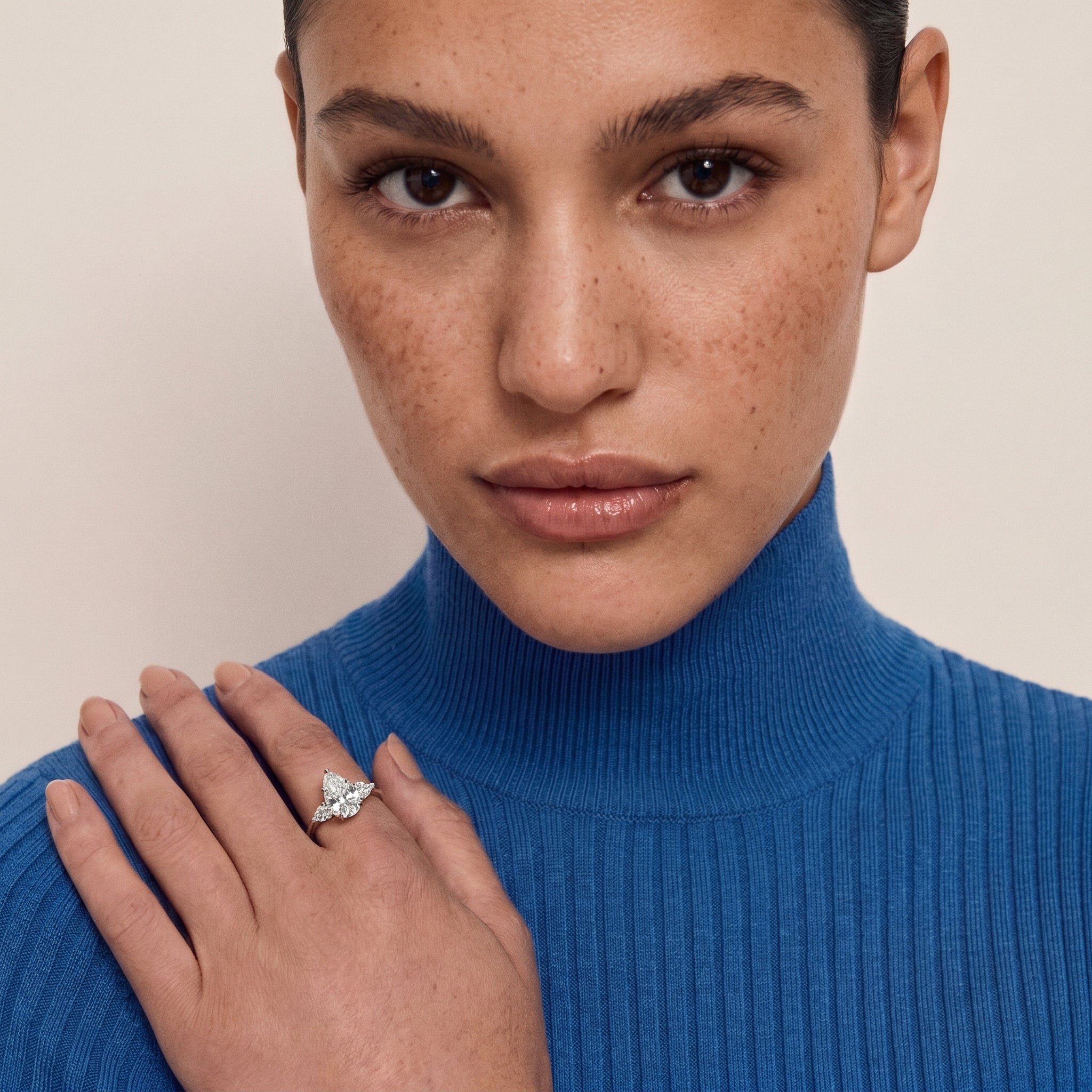 Model wearing the elegant pear cut three-stone cubic zirconia ring on her left ring finger, paired with a sophisticated blue ribbed turtleneck, showcasing the ring's size and refined style on a hand with natural freckles.