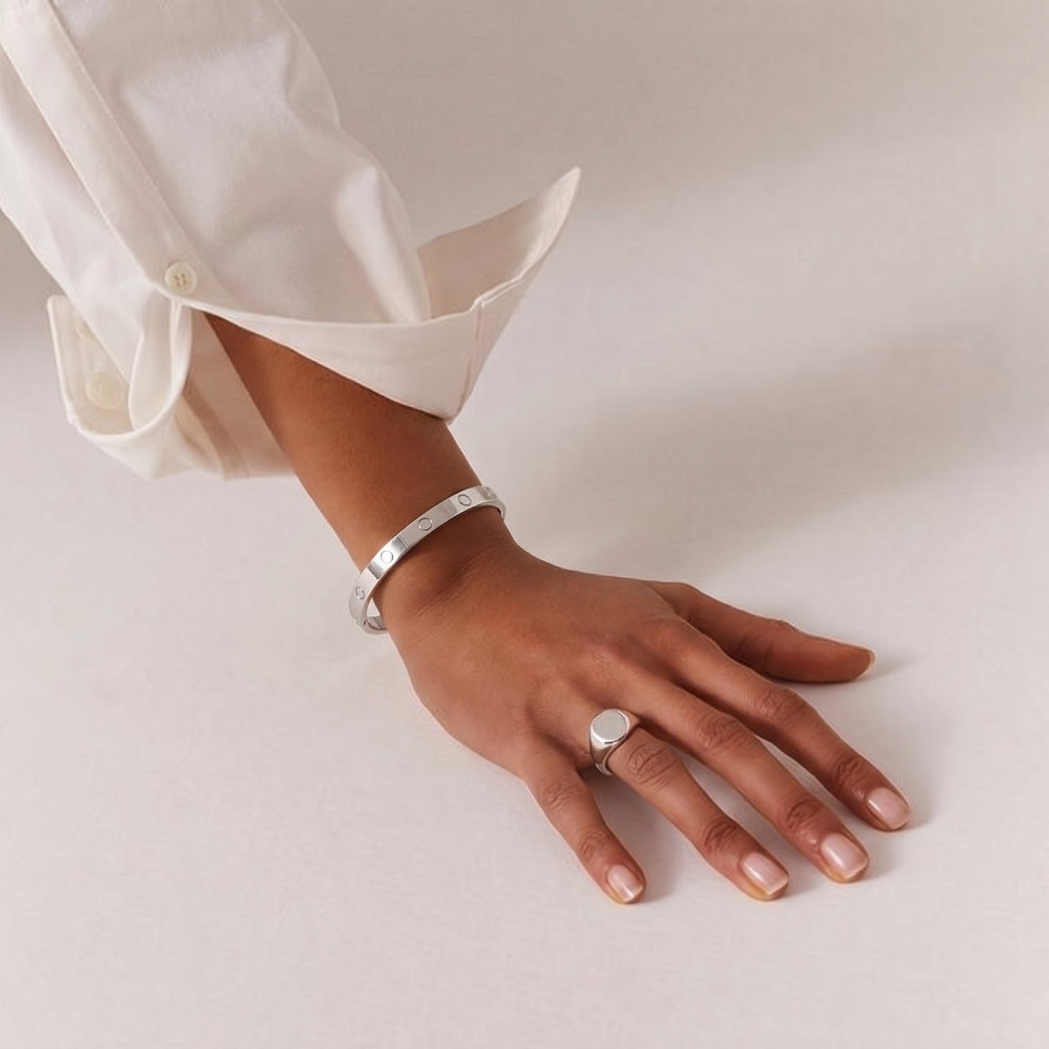 A person's arm wearing a polished silver-toned titanium steel screw-motif bangle, paired with a matching signet ring and a crisp white shirt, showcasing sophisticated everyday style.