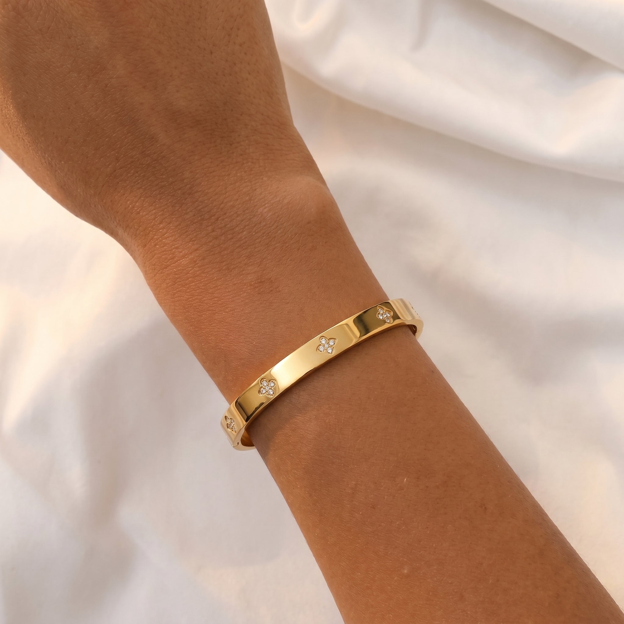 Detail shot of a wrist adorned with the gold-plated titanium steel bangle bracelet, highlighting the brilliant diamond clover motifs against a soft, draped white fabric.