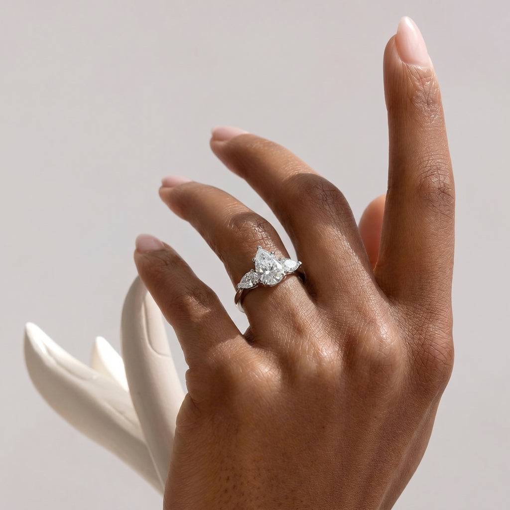 A hand with meticulously manicured nails gracefully displaying the pear cut three-stone cubic zirconia ring on the ring finger, set against a soft, light background with subtle shadows.